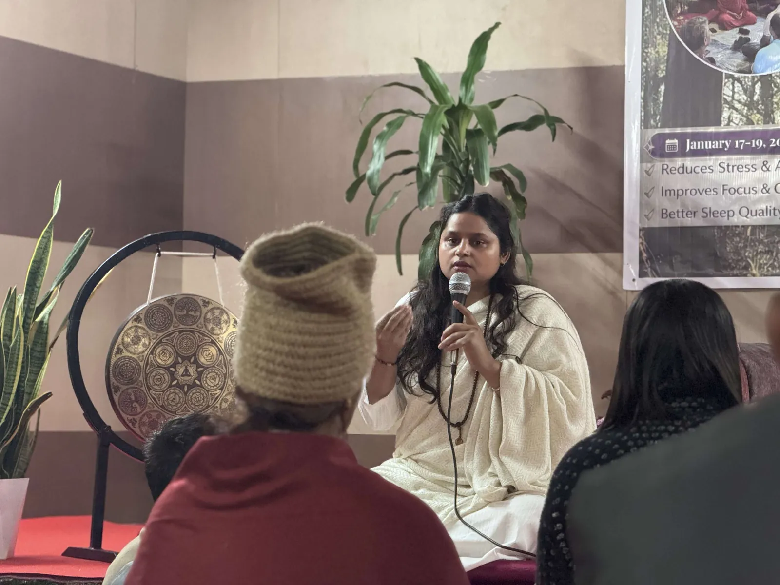A teacher in white robes speaking into a microphone at a public session, seated in front of a large decorated gong with listeners in the foreground