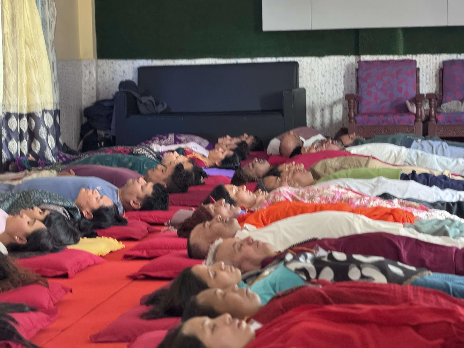 Dozens of participants resting side by side on red matting with their eyes closed, wrapped in shawls and blankets