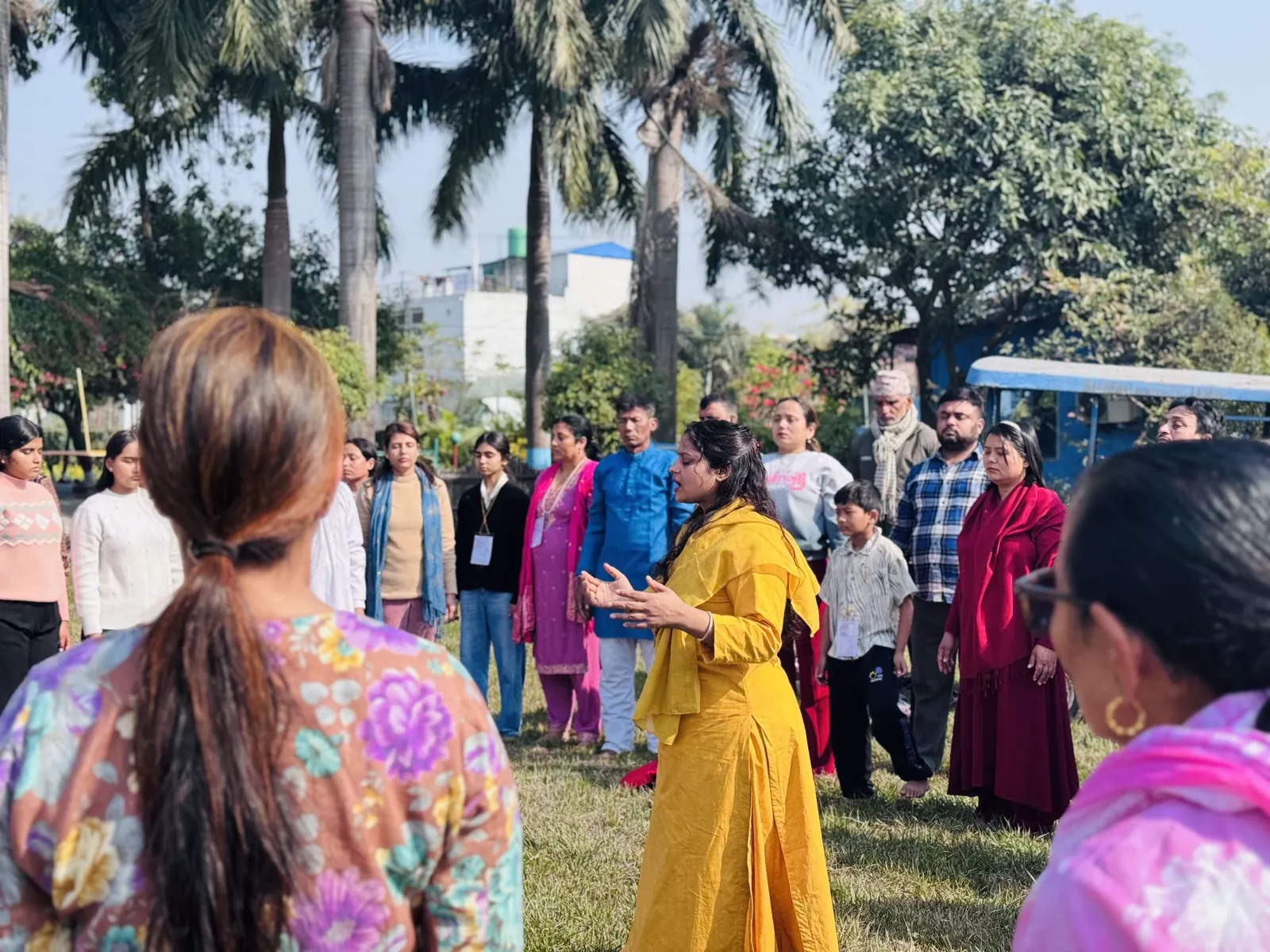A teacher in a yellow kurta guiding a standing group through a practice on a sunlit lawn under palm trees
