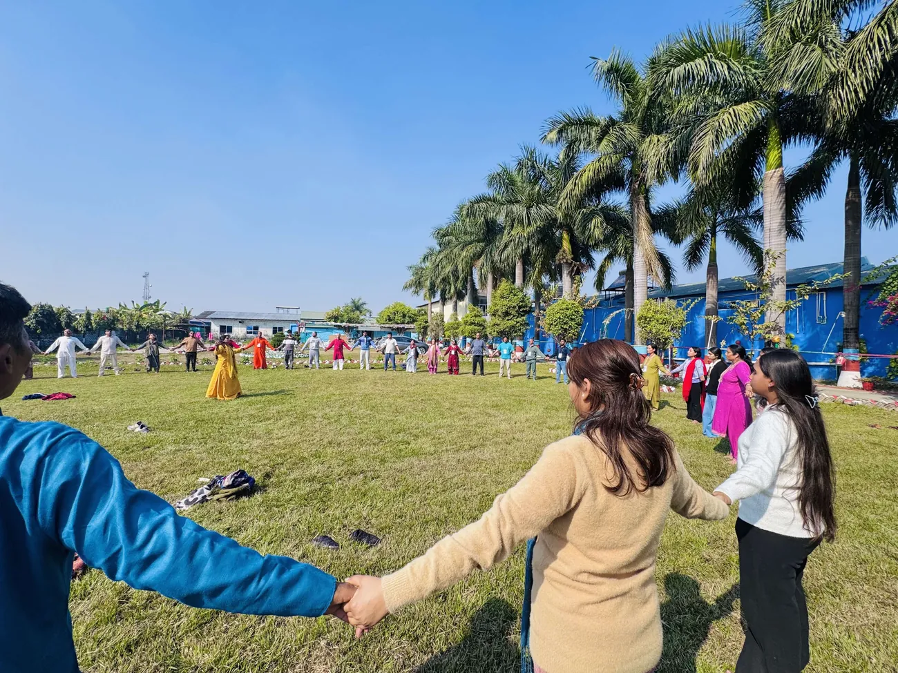 Participants holding hands in a circle across a sunlit lawn, seen from inside the circle, with palm trees along one side