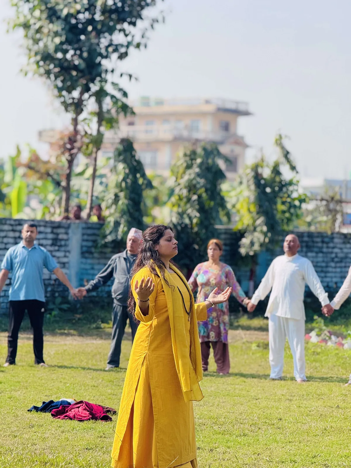 A teacher standing with open palms and eyes closed while the group behind her holds hands in a circle on the grass