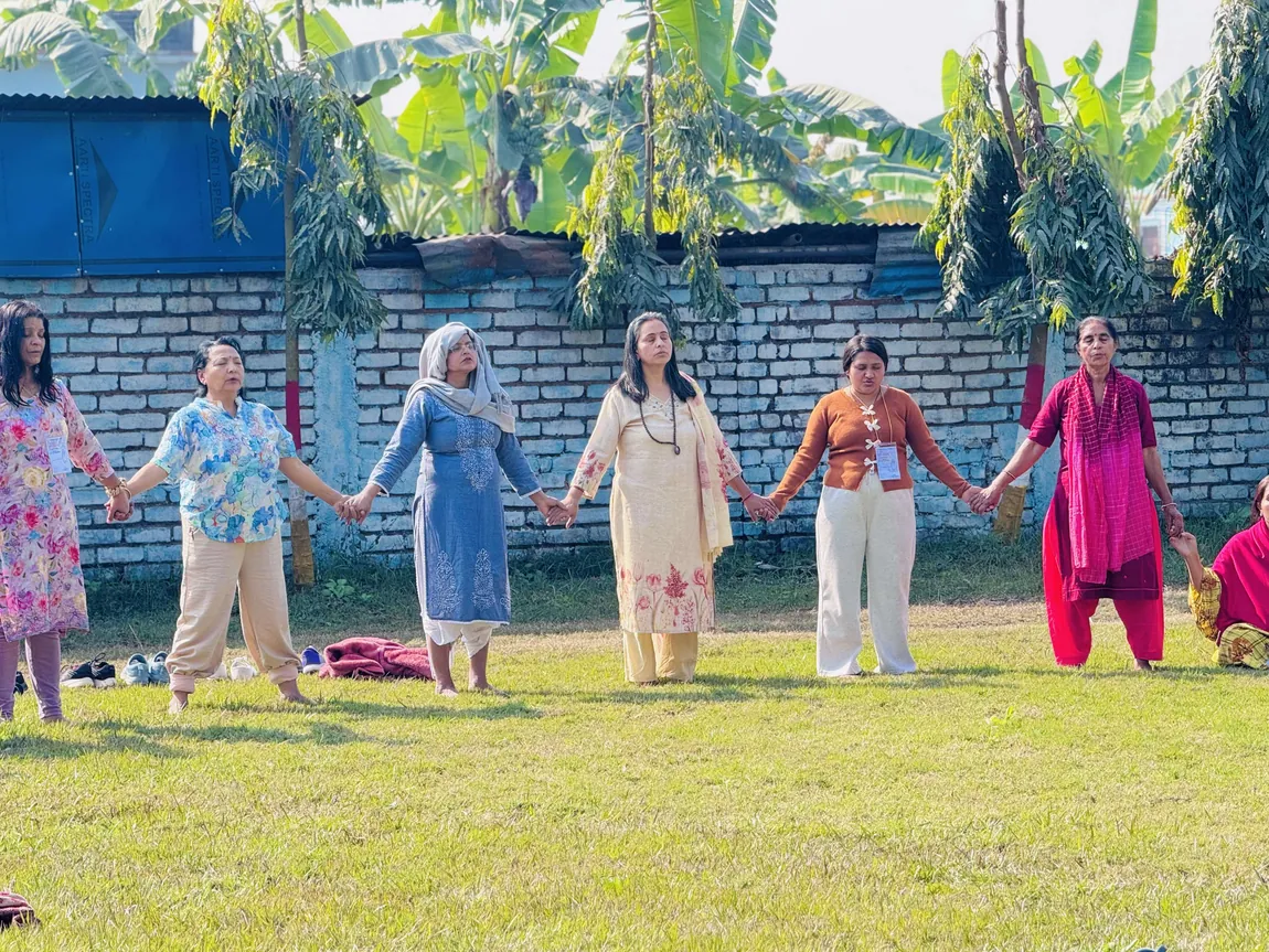 Six women standing hand in hand with their eyes closed in front of a brick wall and banana trees during an outdoor practice