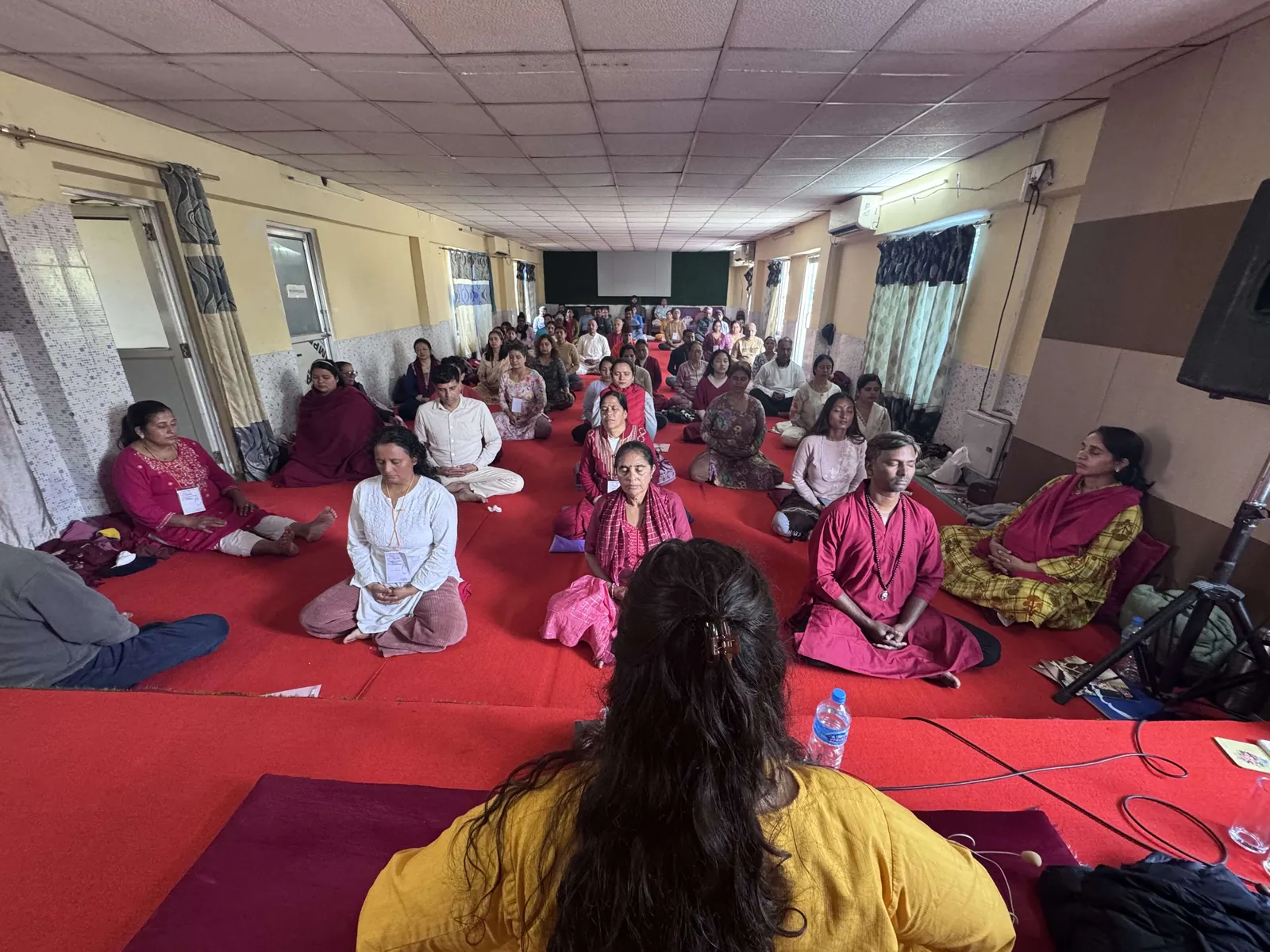A hall full of people sitting cross-legged in meditation on red matting, eyes closed, led by a teacher seated with her back to the camera