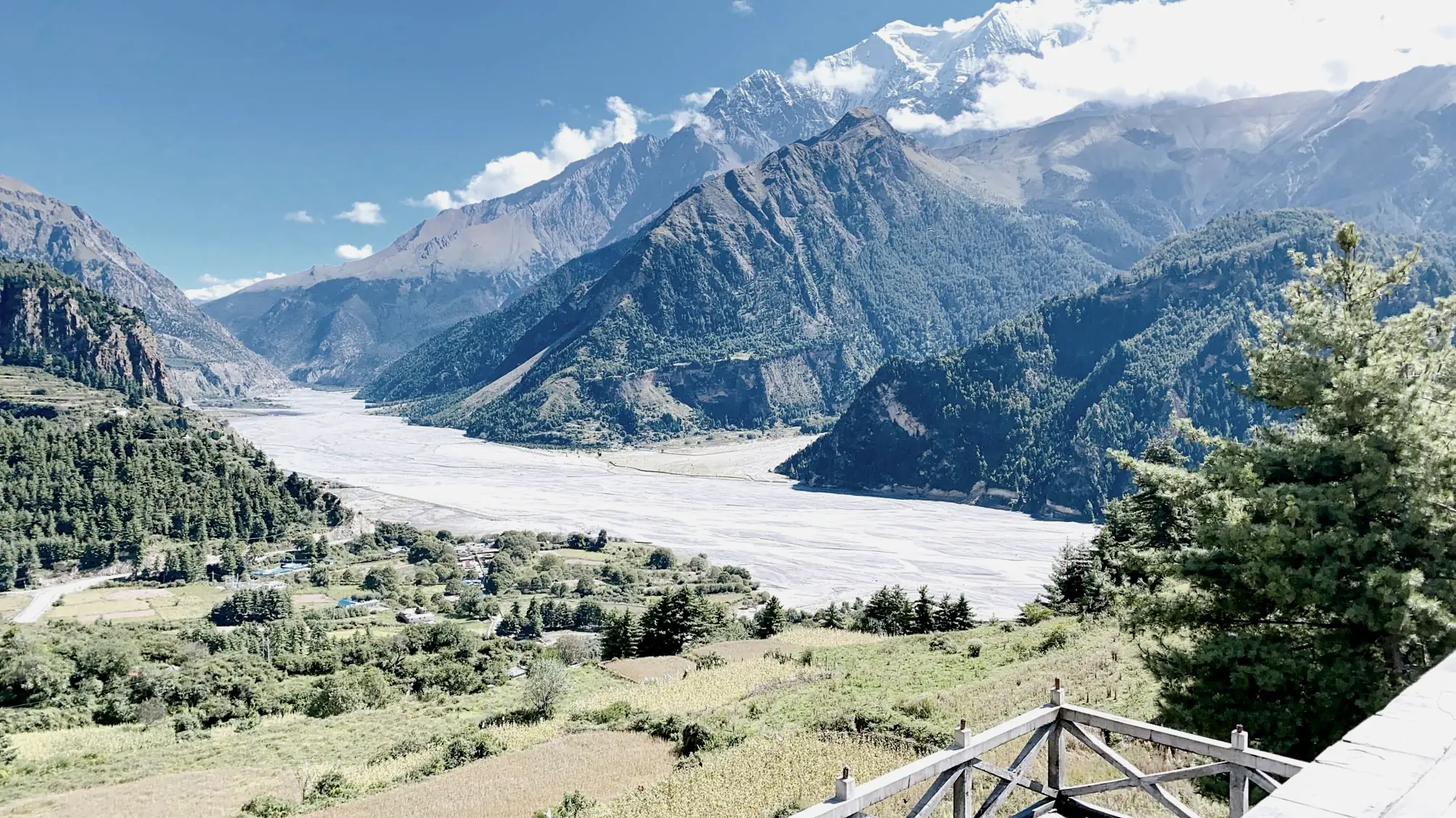The wide, pale riverbed of the Kali Gandaki winding between forested ridges below a timber railing, with a snow peak in cloud above