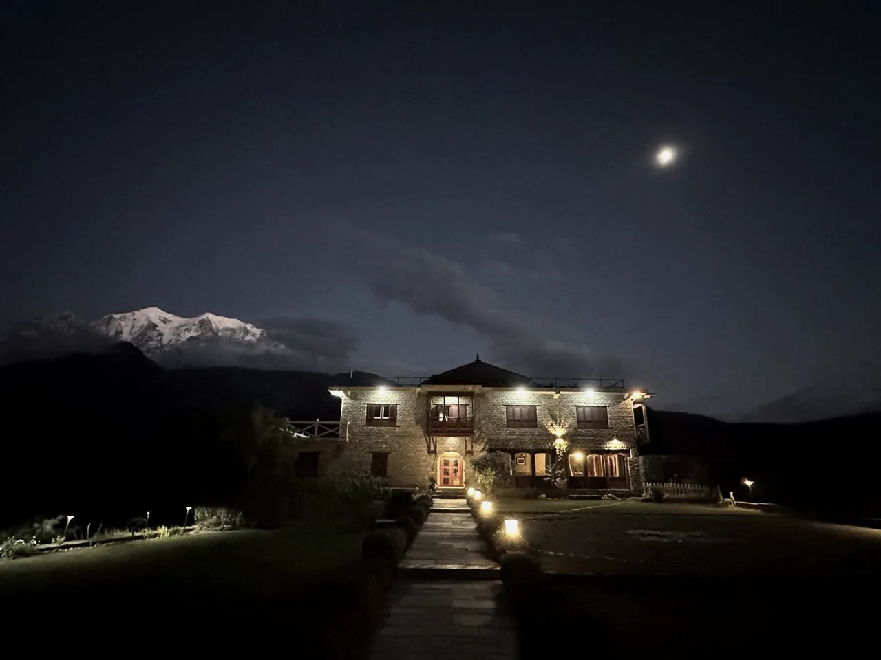 Lodge Thasang Village lit at night under a bright moon, lamps along the stone path to its door and a pale snow peak in the dark to the left
