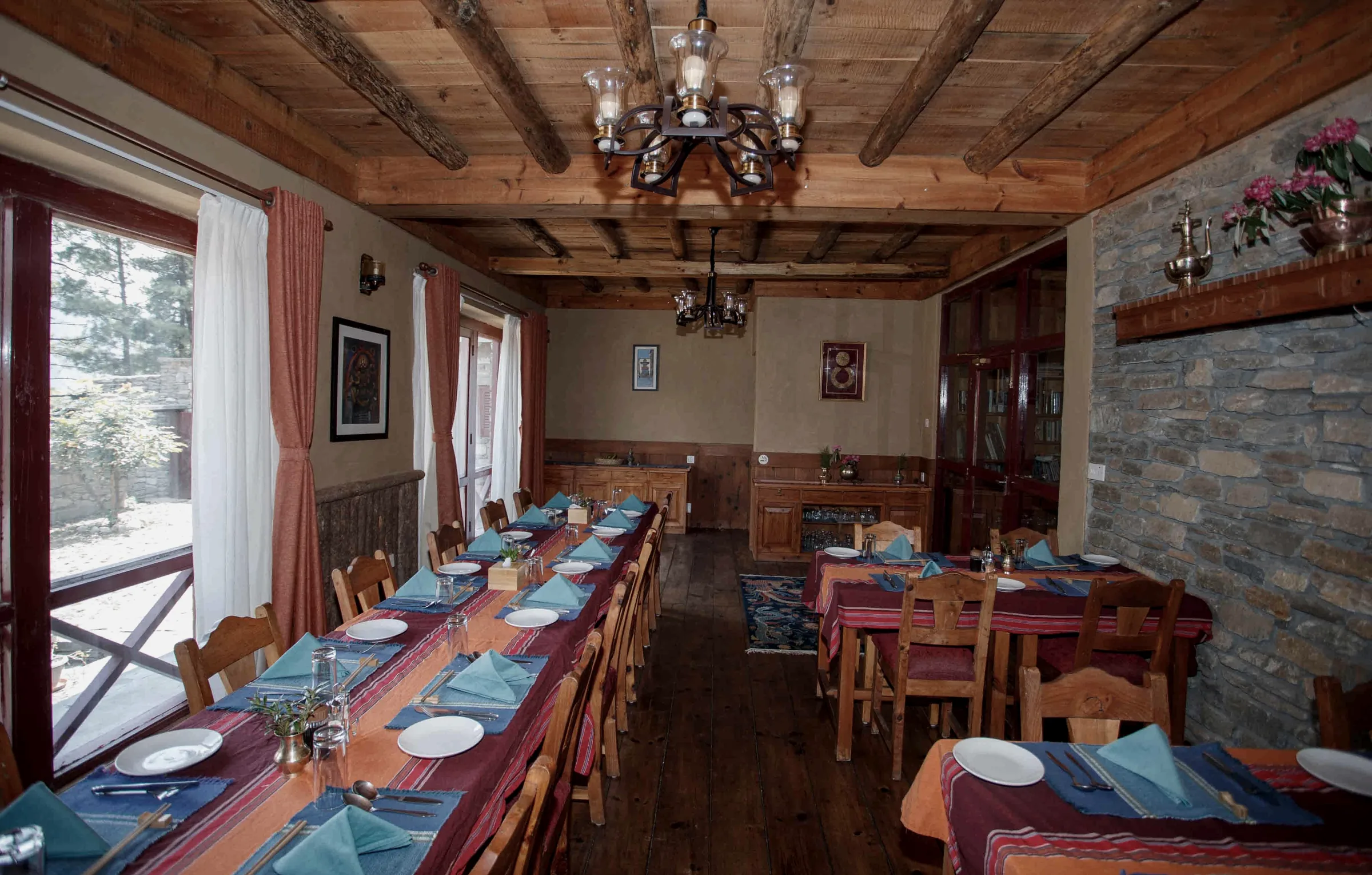 A long dining table laid with blue napkins, plates and striped runners beside tall curtained windows, with a stone wall and smaller tables across the room