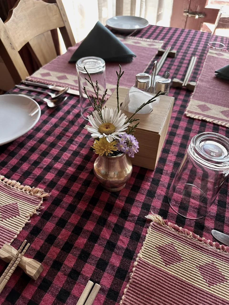 A small brass vase of wildflowers on a checked tablecloth, set with upturned glasses, plates and chopsticks