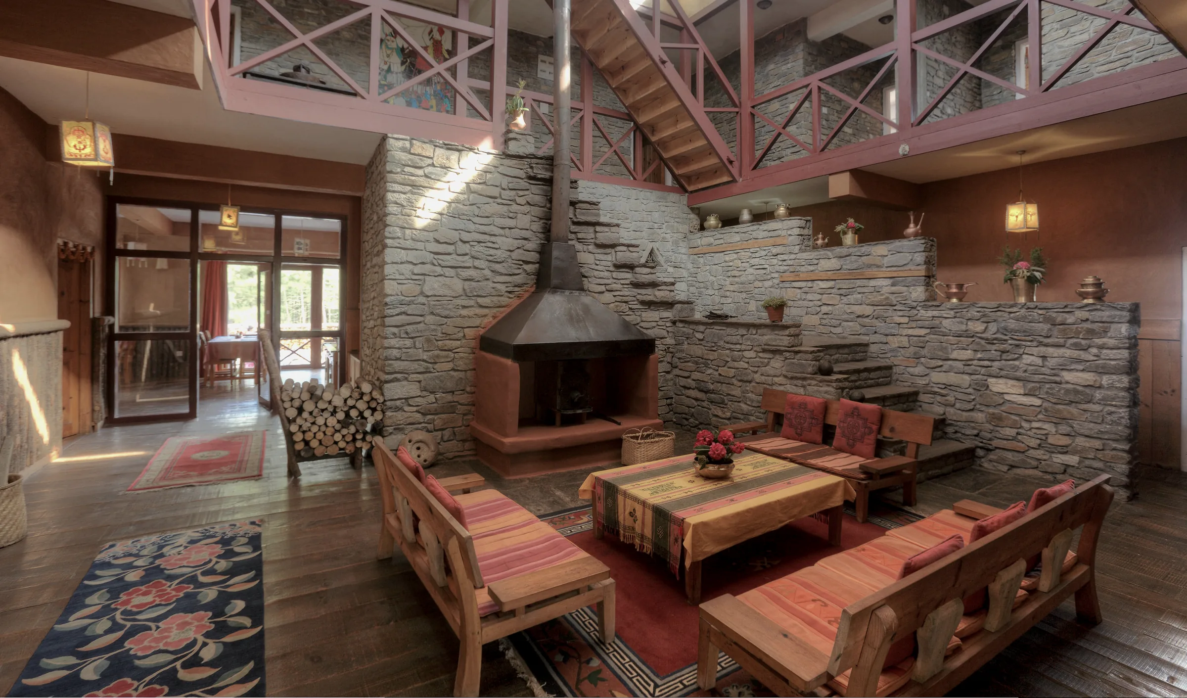 The double-height lobby: a hooded fireplace set into a stone wall, stacked firewood, timber benches around a low table, and a gallery and staircase above