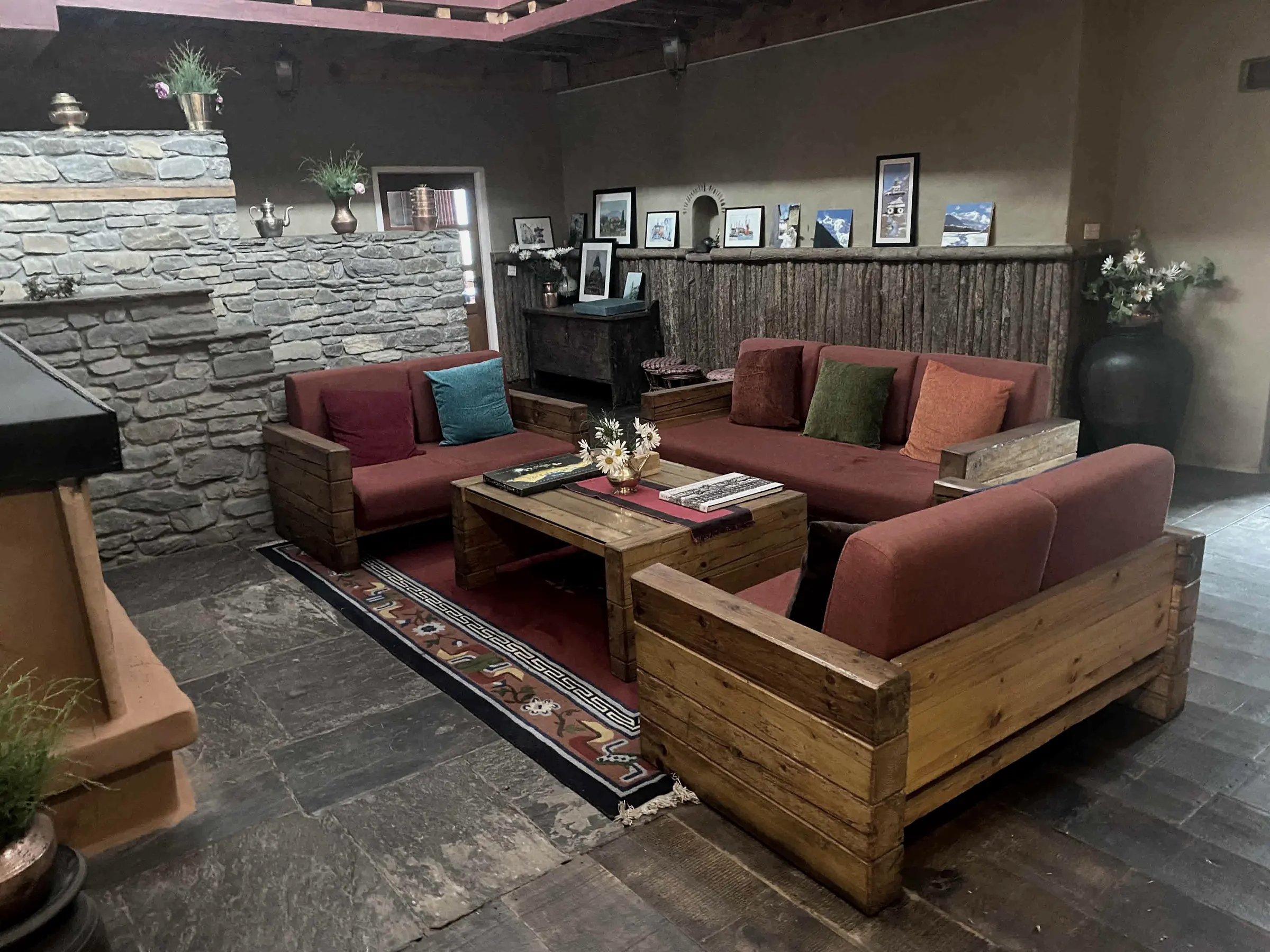 Timber-framed sofas with red cushions around a low wooden table on a patterned rug, with a stone wall and a row of framed mountain pictures behind
