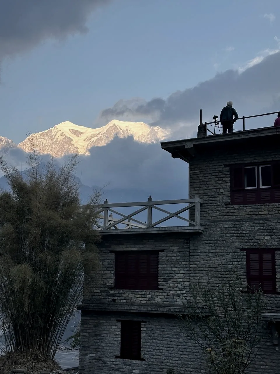 A snow peak lit gold above cloud beyond the lodge's stone walls, with a figure standing at the rooftop railing