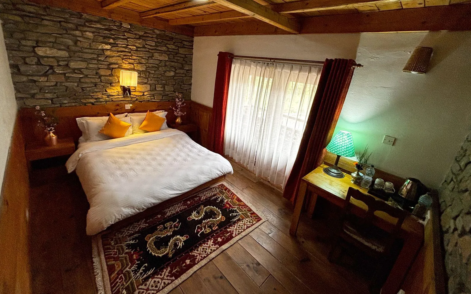 A single deluxe room: one bed with a white duvet and two mustard cushions against a lamplit stone wall, a dragon rug on the plank floor, and a desk with a green lamp and kettle beside a curtained window