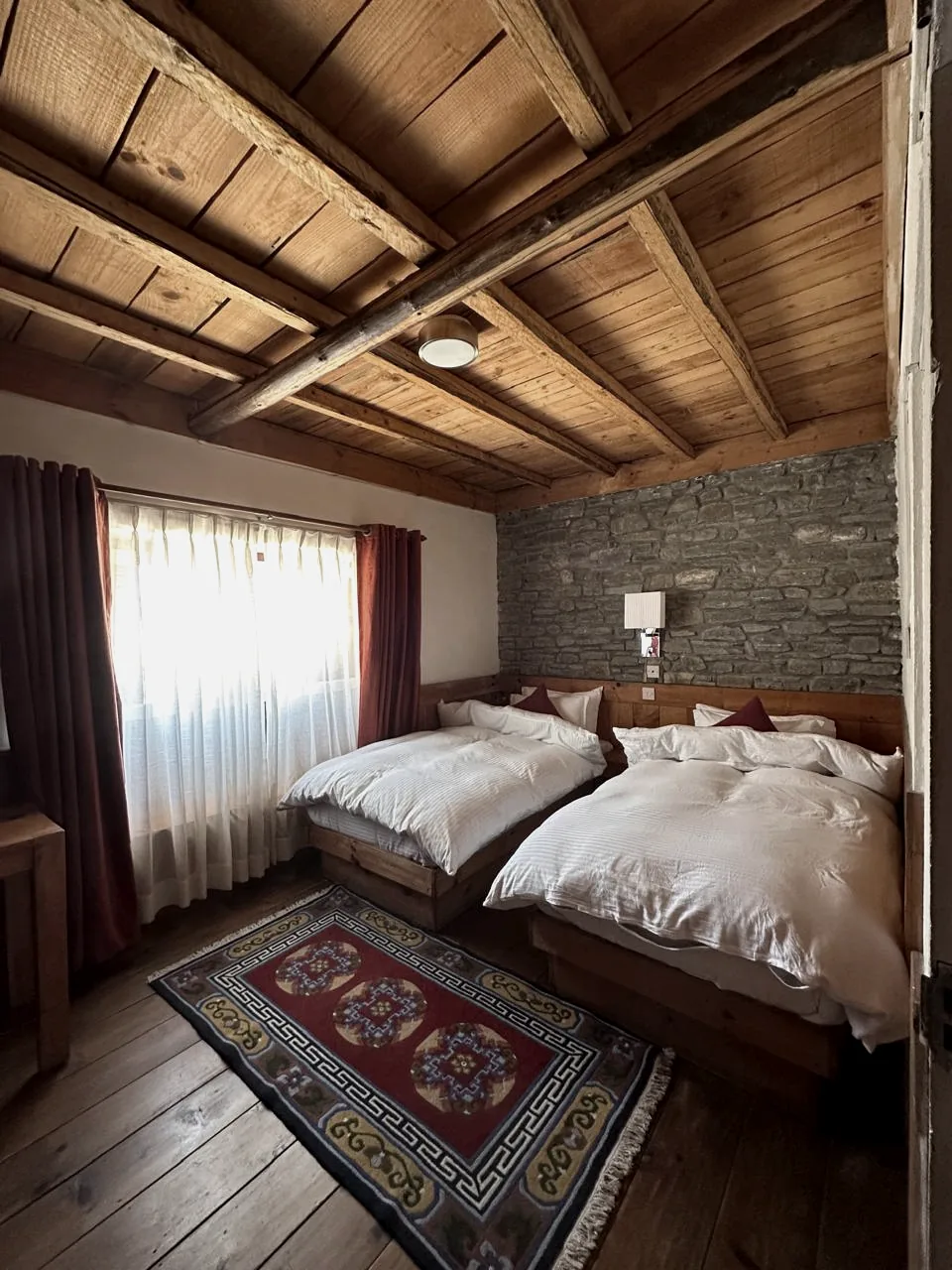 Two beds made up with white duvets against a grey stone wall, under a planked timber ceiling, with a patterned red rug and a sunlit curtained window
