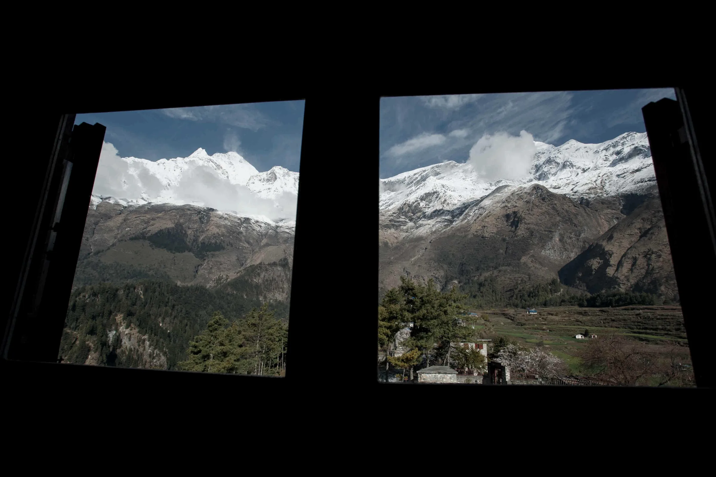 Snow-covered peaks above forested slopes and a green valley floor, framed by two open windows in a darkened room