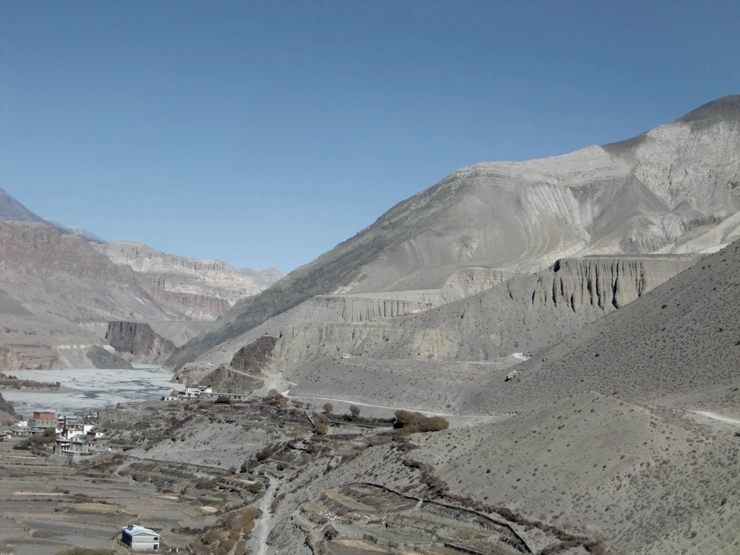 Bare, wind-carved cliffs and grey slopes above a braided riverbed and a small terraced village, under a cloudless sky
