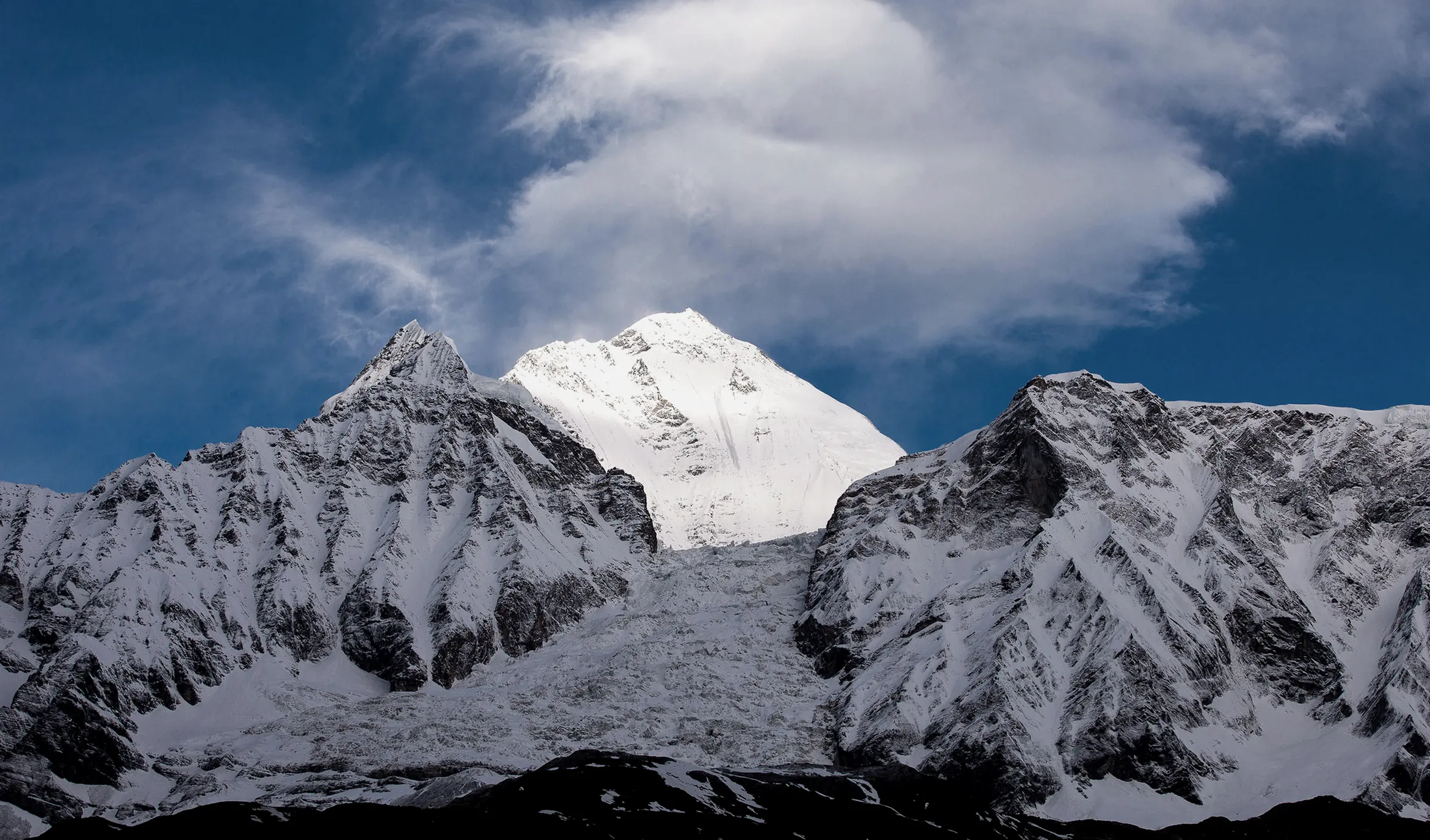 A snow summit rising behind two dark, snow-streaked ridges, with a sweep of white cloud across a blue sky