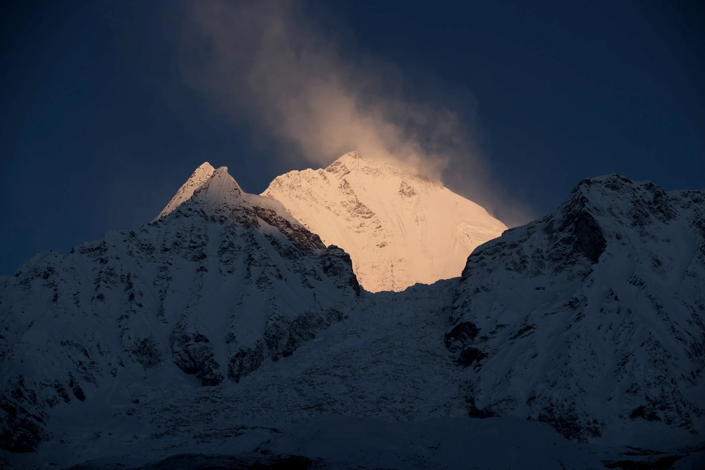 A snow summit lit by low sun against a dark blue sky, a plume of cloud streaming from its top