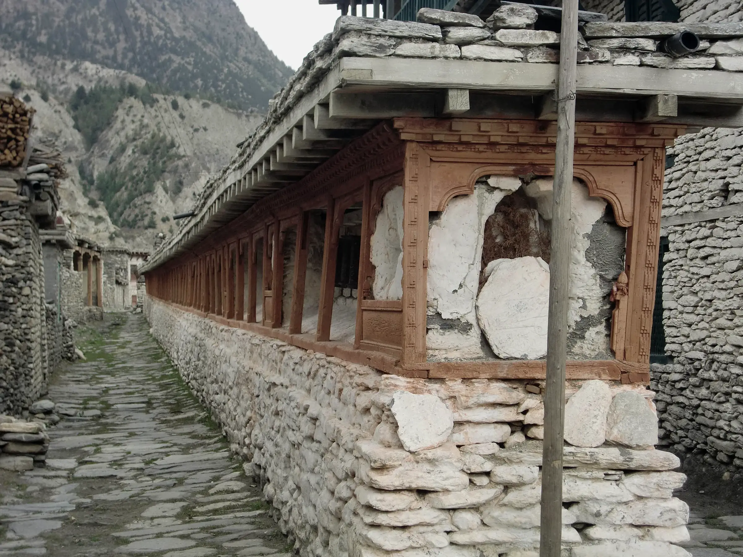 A long roofed wall of carved timber niches on a whitewashed stone base, beside a stone-paved village lane