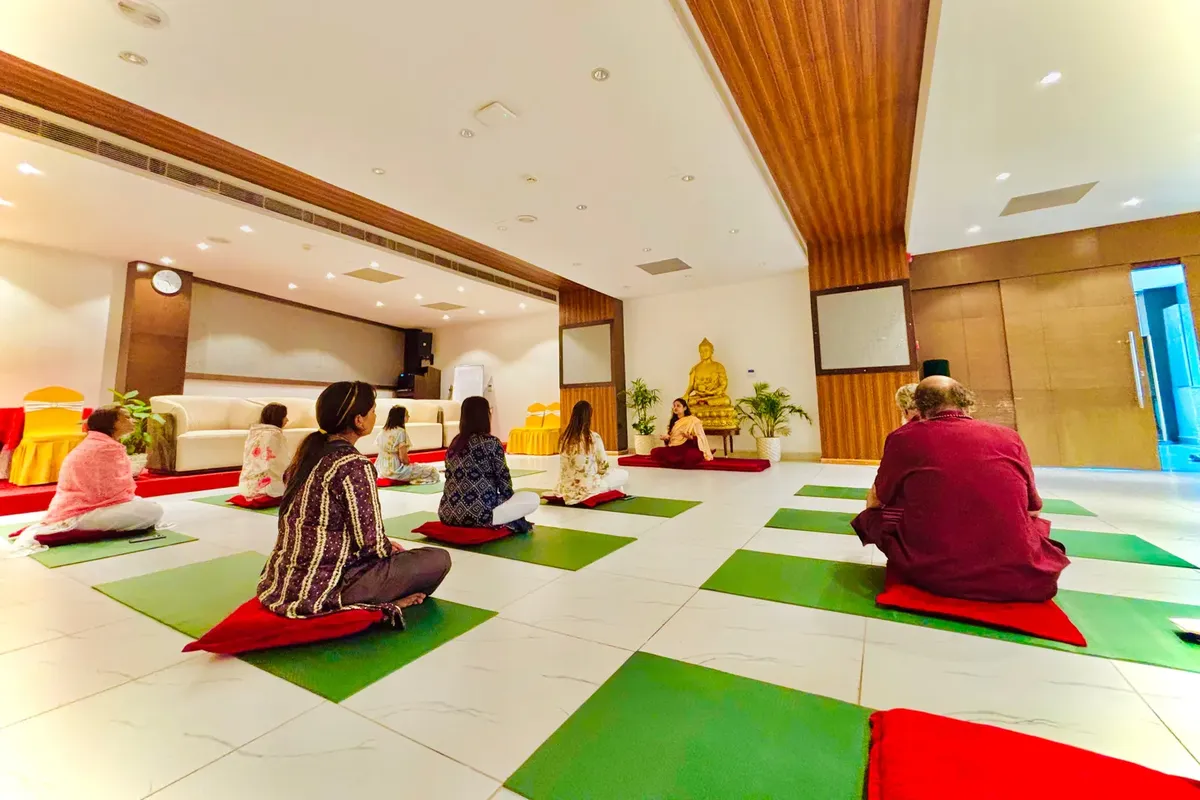 Students seated on mats facing their teacher in a bright yoga hall with a golden Buddha statue