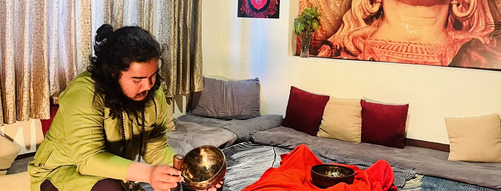 A practitioner holding a hammered metal singing bowl beside a guest resting under a red blanket