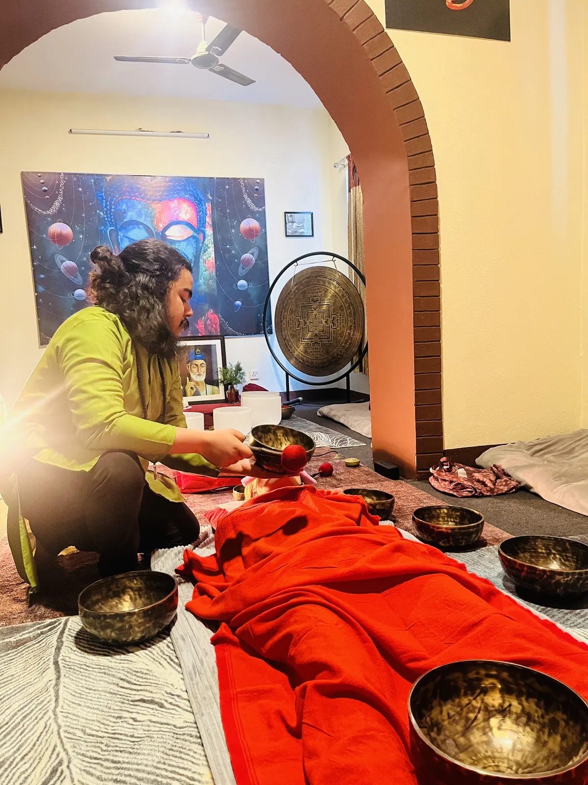 A practitioner plays a bowl beside a guest resting under a red blanket, with more bowls set along the guest and crystal bowls and a gong through the archway