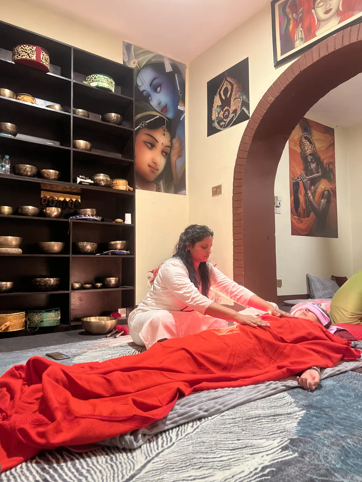 A practitioner giving hands-on energy healing to a resting guest, shelves of singing bowls behind